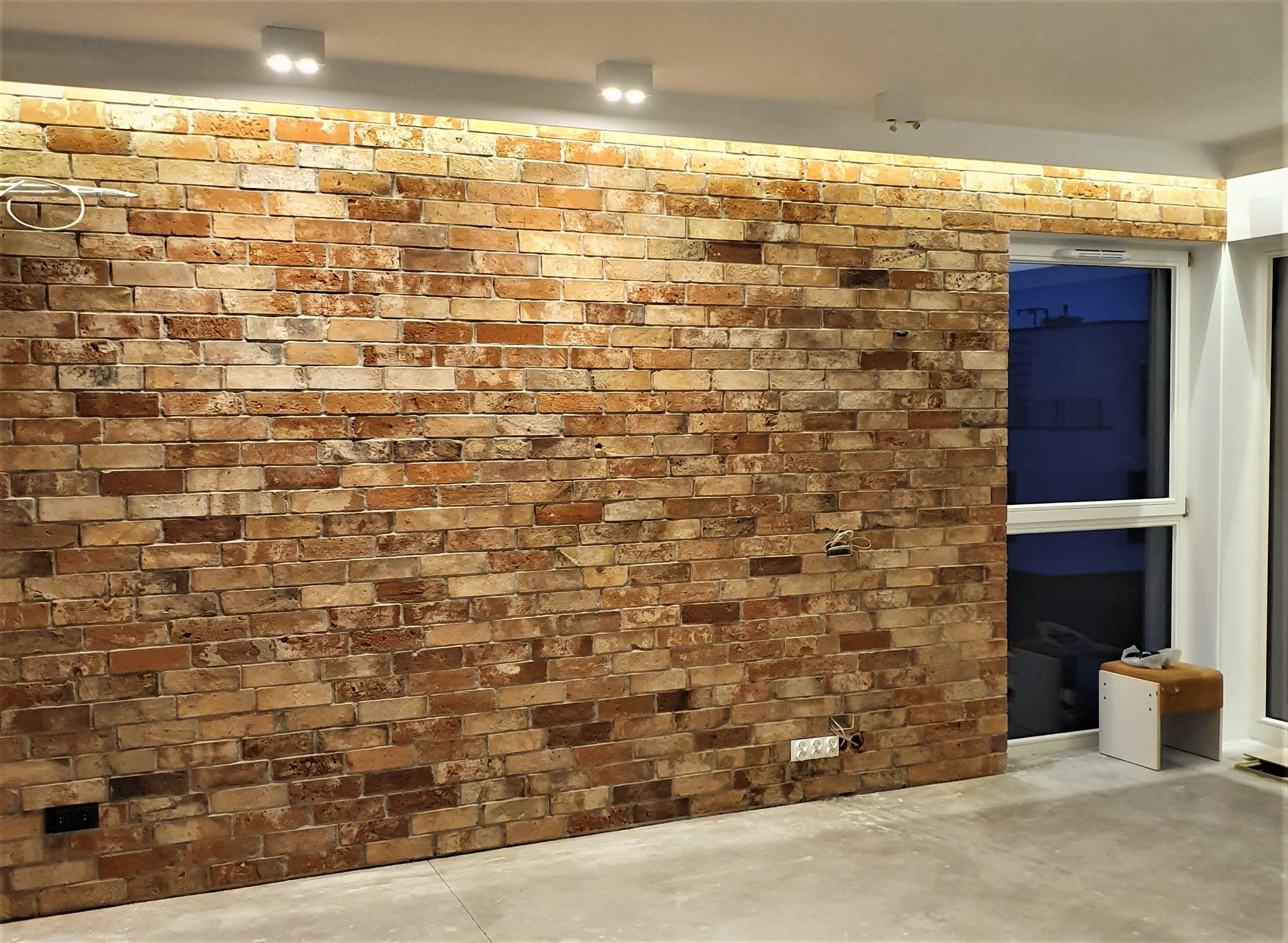 Ściana z cegły klinkierowej w odcieniach brązu i beżu, oświetlona punktowymi lampami sufitowymi i listwą LED, widoczne gniazdka elektryczne, w tle okno z widokiem na budynek po zmroku i mały taboret.