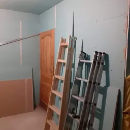 Wnętrze w trakcie montażu płyt kartonowo-gipsowych na ścianach i suficie, widoczne dwie drabiny opierające się o ścianę, oraz częściowo widoczne drzwi drewniane.