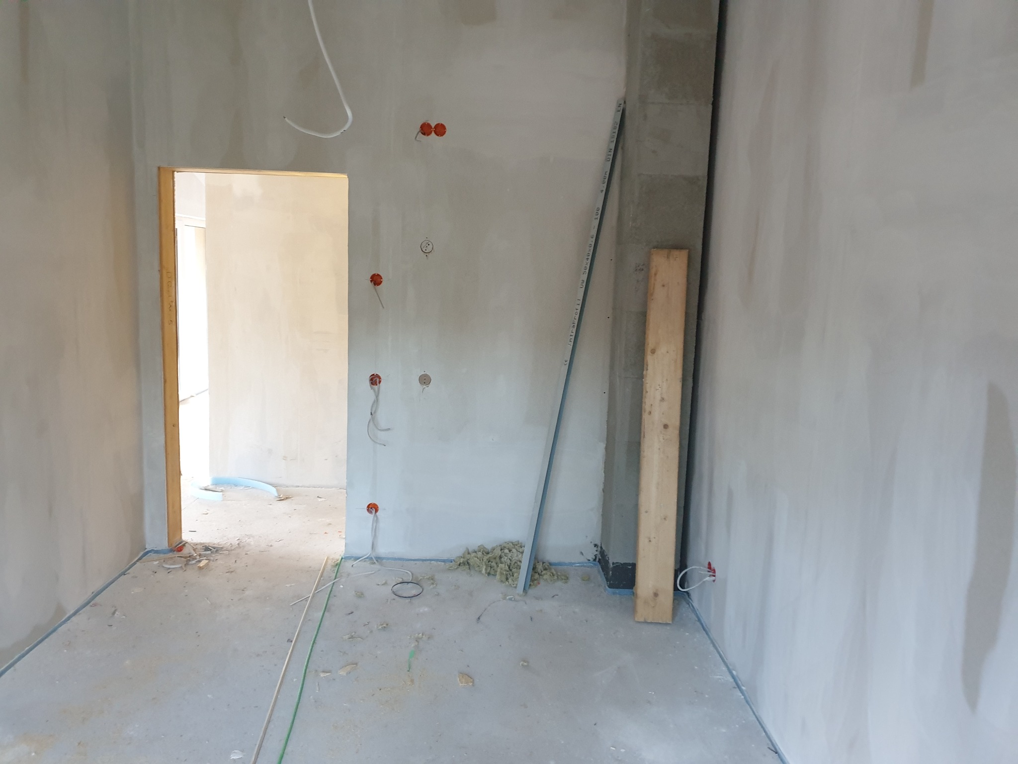 Surowe wnętrze w trakcie prac: ściany po gładzi, widoczne punkty elektryczne, wystające przewody, fragmenty wełny mineralnej na podłodze, listwa przypodłogowa w kolorze niebieskim, ościeżnica...