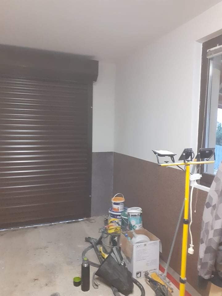 Wnętrze garażu w trakcie malowania: widoczna brązowa brama garażowa, pomalowana górna część ściany na biało i dolna na brązowo, puszki z farbą oraz lampa halogenowa na żółtym statywie.