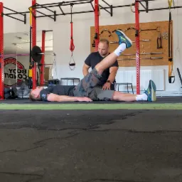 Mężczyzna leżący na podłodze w siłowni, podnoszący nogę do góry, podczas gdy druga osoba przytrzymuje mu kolano, w tle widoczne wyposażenie siłowni i logo.