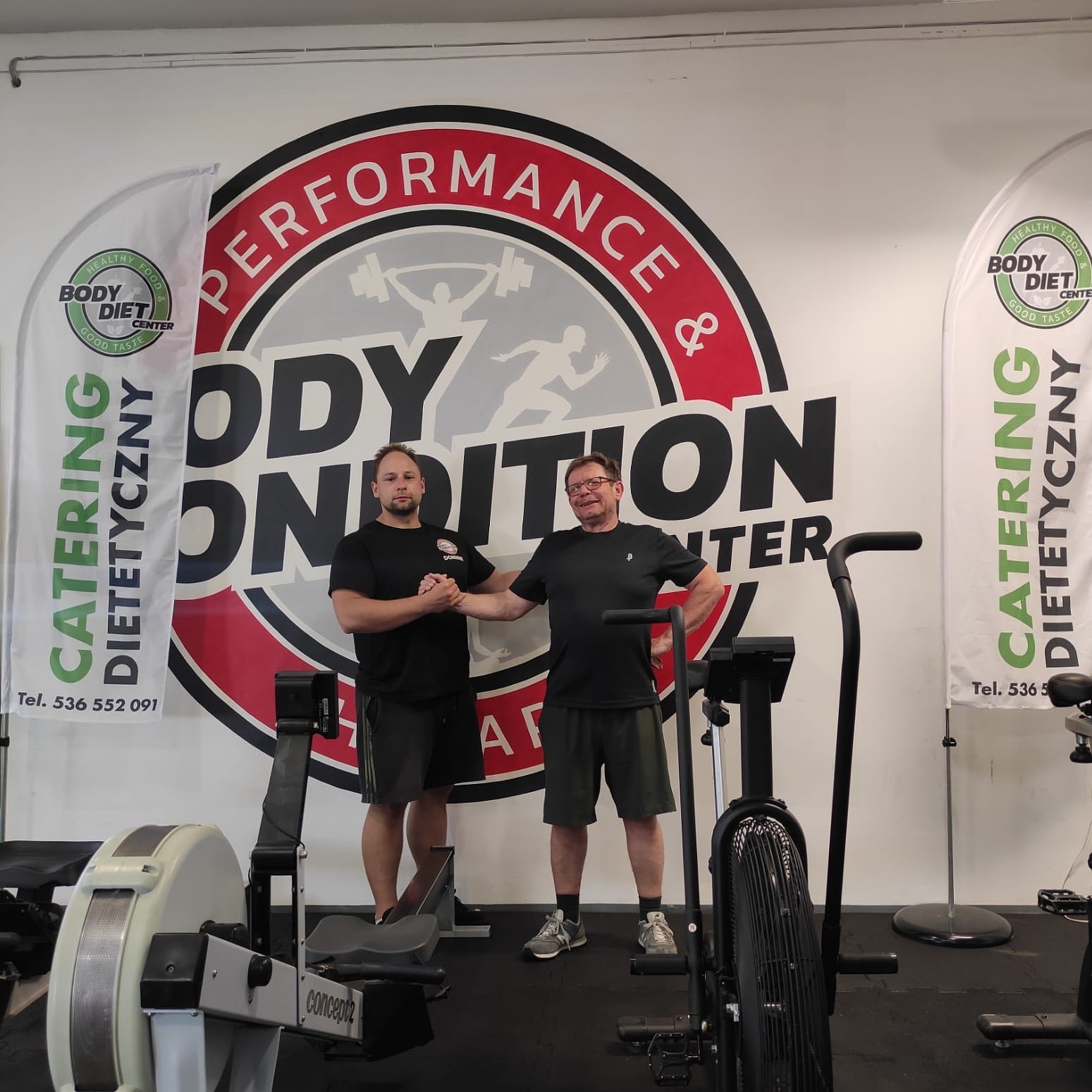 Mężczyzna w czarnej koszulce ściska dłoń drugiemu mężczyźnie na tle logotypu firmy Body Condition Center i sprzętu do ćwiczeń, widoczne flagi reklamowe cateringu dietetycznego.
