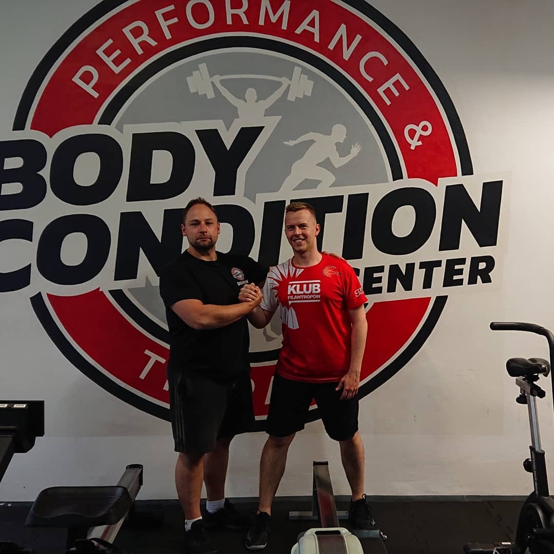 Dwóch mężczyzn w sportowych strojach, ściskających dłonie na tle logo siłowni Body Condition Center, z widocznym sprzętem do ćwiczeń w tle.