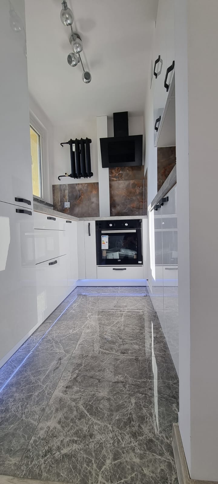 Nowoczesna kuchnia z białymi, lakierowanymi szafkami, czarnymi uchwytami i marmurową podłogą z podświetleniem LED, widoczny czarny okap i grzejnik żeberkowy.