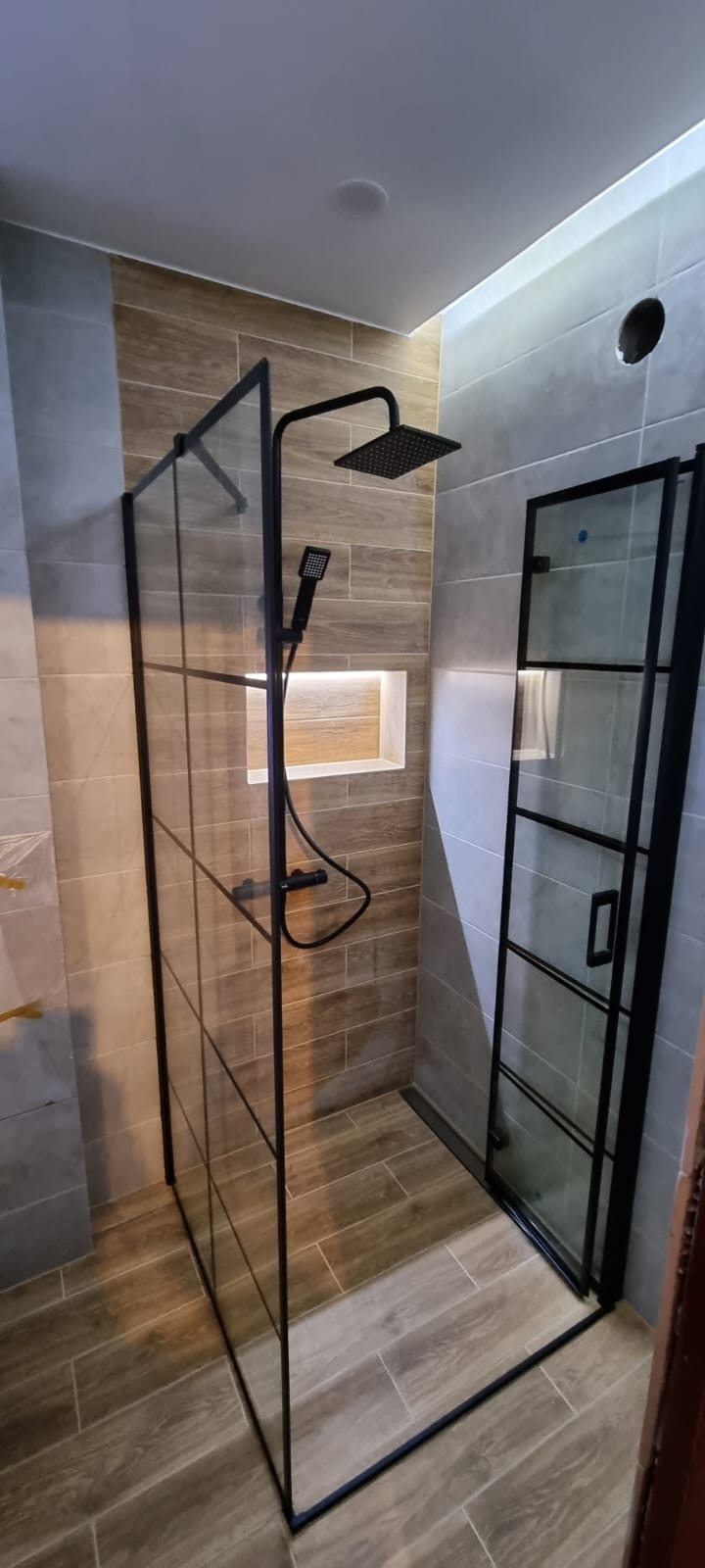 Nowoczesna kabina prysznicowa z czarnymi profilami i deszczownicą w wykończonej łazience z płytkami imitującymi drewno i szarymi płytkami na ścianach.