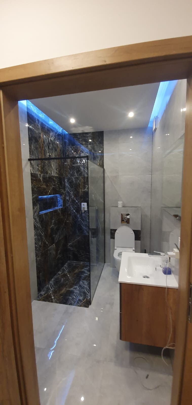 Nowoczesna łazienka z kabiną prysznicową wykończoną czarnym marmurem z podświetleniem LED, umywalką z szafką z jasnego drewna oraz toaletą w odcieniach bieli i szarości, widok zza drewnianej...