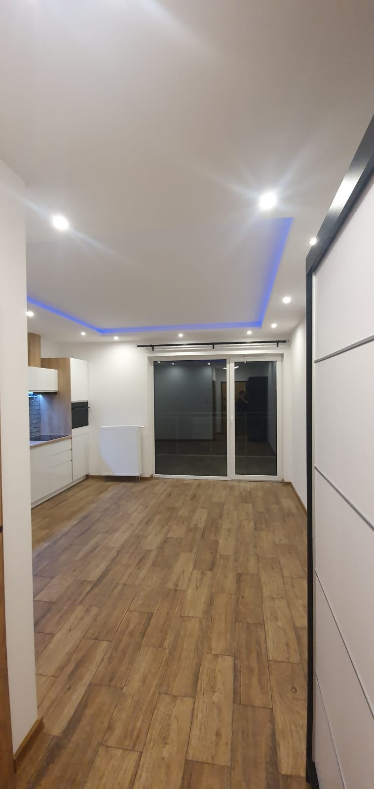 Nowoczesne wnętrze mieszkania z drewnianą podłogą, białymi ścianami, przesuwnymi drzwiami balkonowymi i oświetleniem LED w suficie.