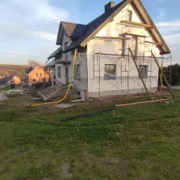 Dom jednorodzinny w trakcie ocieplania elewacji, widoczne rusztowania, styropian na ścianach i przygotowane okna, w tle inne budynki i błękitne niebo.