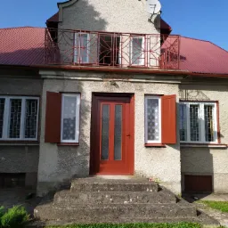 Dwukondygnacyjny dom z czerwonym dachem i balkonem z dekoracyjną balustradą, czerwone okiennice częściowo zasłaniają okna, wejście poprzedzone betonowymi schodami.