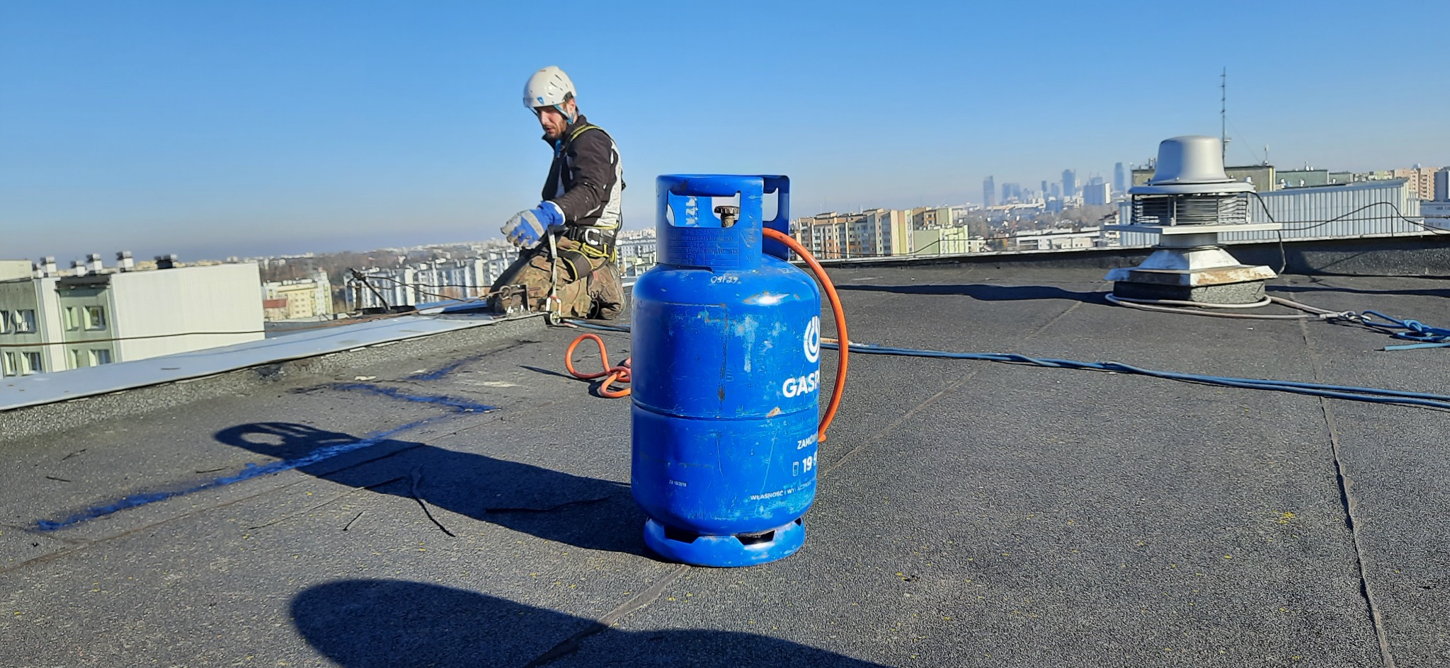 Pracownik w kasku na dachu z butlą gazową na tle panoramy miasta. Widoczne zabezpieczenia linowe. Praca na wysokości, słoneczny dzień.