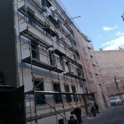 STAHLER Patryk Pobłocki - Wielopiętrowy budynek w trakcie ocieplania, osłonięty rusztowaniami i siatką ochronną z widocznymi pracownikami na różnych poziomach oraz zaparkowanym samochodem w tle.