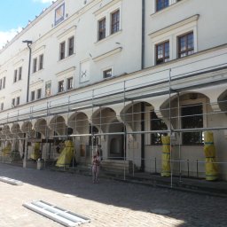 STAHLER Patryk Pobłocki - Biały budynek z arkadami w trakcie prac renowacyjnych, osłonięte filary, rusztowania, kobieta z dzieckiem w tle