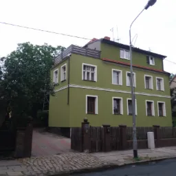 Odnowiony, dwupiętrowy dom z elewacją w odcieniach zieleni, z białymi ramami okiennymi i ciemnobrązową podmurówką, widoczny z poziomu ulicy w pochmurny dzień.