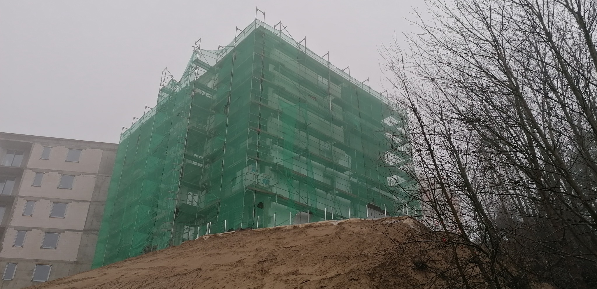 Wielopiętrowy budynek w trakcie prac elewacyjnych, osłonięty zieloną siatką rusztowaniową, widoczny w mglisty dzień, z hałdą ziemi u podstawy i drzewami w tle.