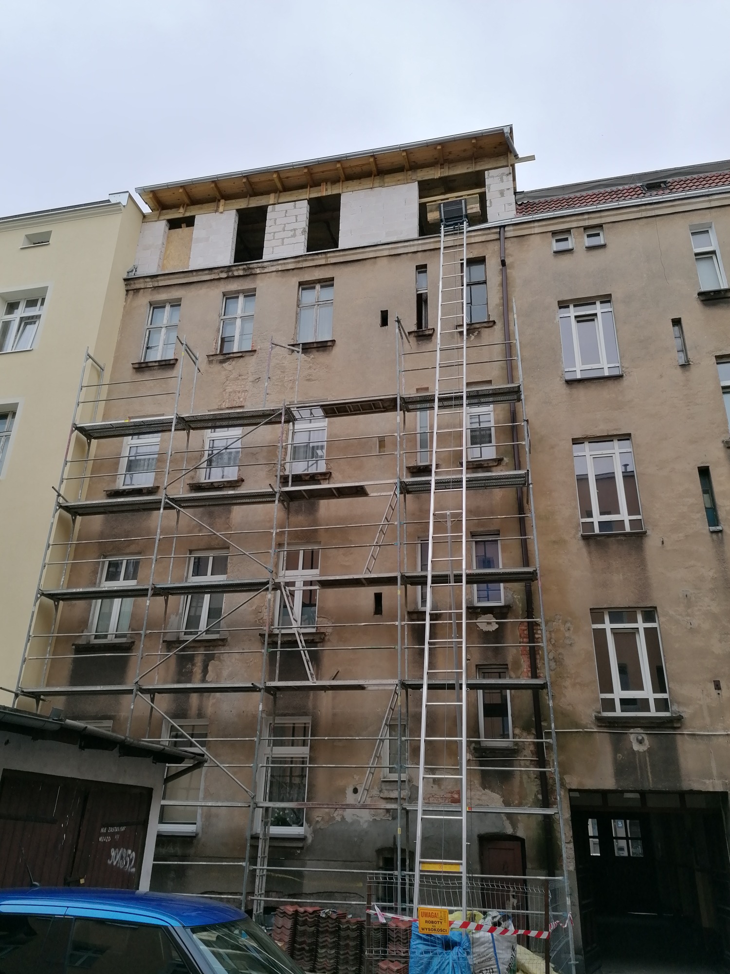 Stare, zaniedbane mieszkanie w trakcie remontu elewacji z rusztowaniem i schodami, widoczne nowe okna i nadbudowa z pustaków.
