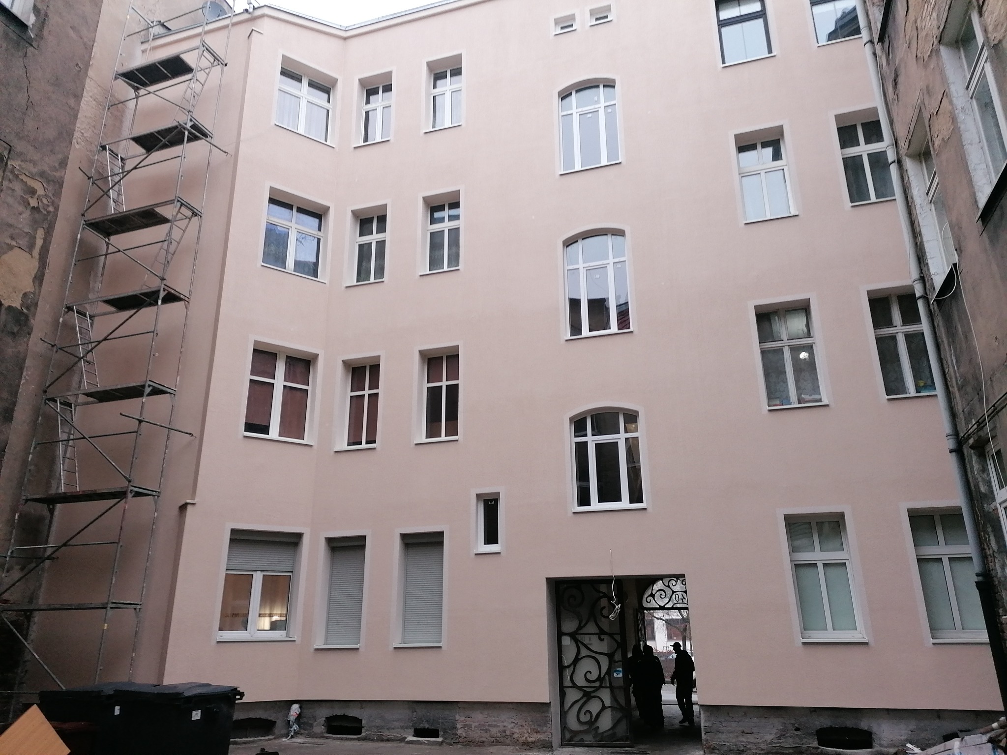 Odnowiona elewacja budynku z oknami o różnych kształtach i rusztowaniem po lewej stronie, widoczne wejście z ozdobną bramą i sylwetki osób w przejściu.