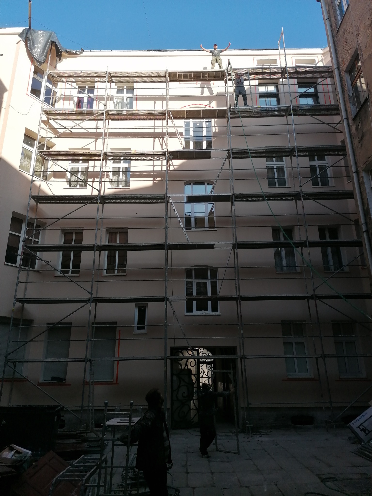 Prace na rusztowaniu przy elewacji kamienicy w trakcie dnia, dwóch pracowników na górze, jeden z rękami w górze, budynek w trakcie renowacji.