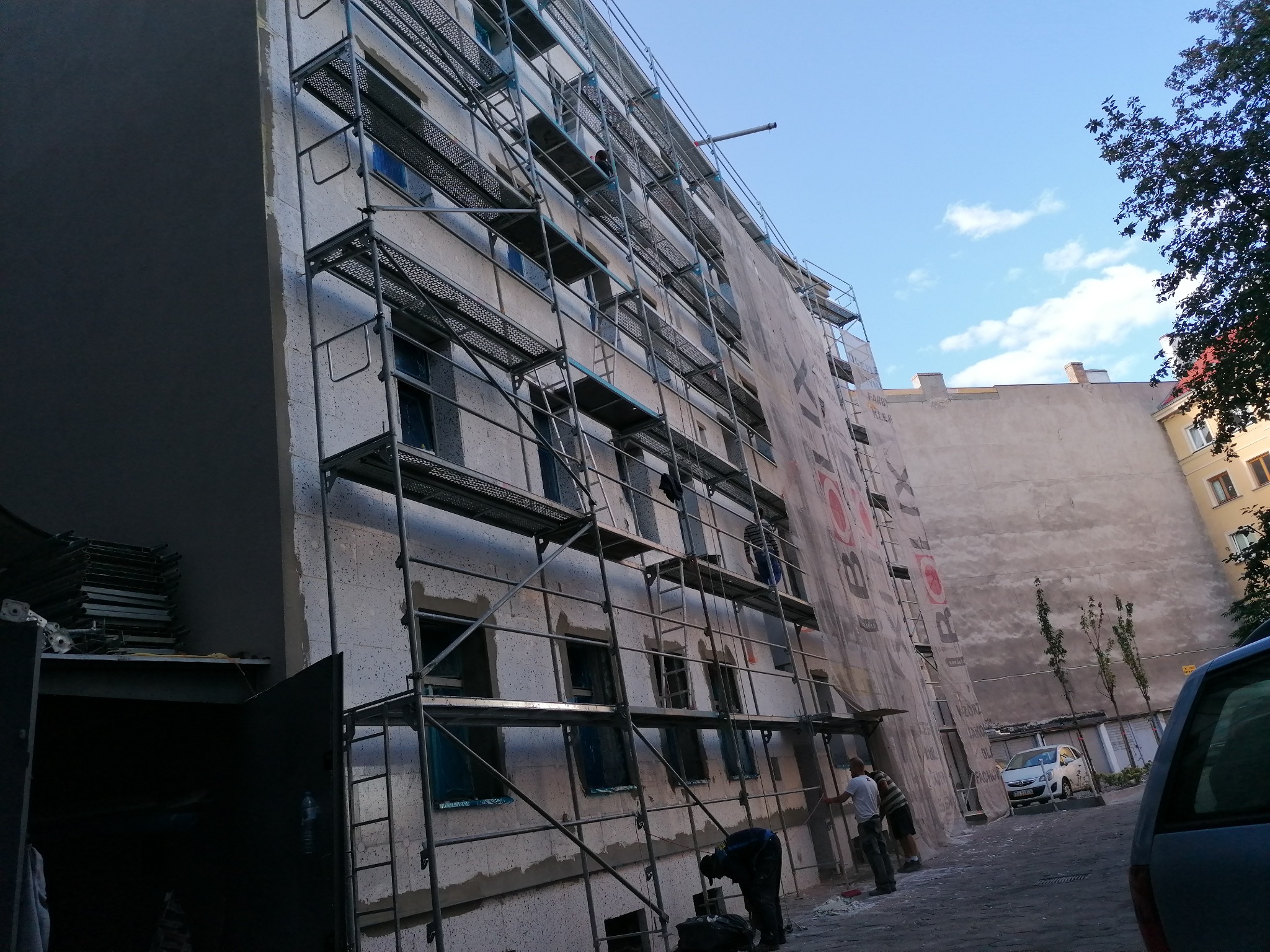Wielopiętrowy budynek w trakcie ocieplania, osłonięty rusztowaniami i siatką ochronną z widocznymi pracownikami na różnych poziomach oraz zaparkowanym samochodem w tle.