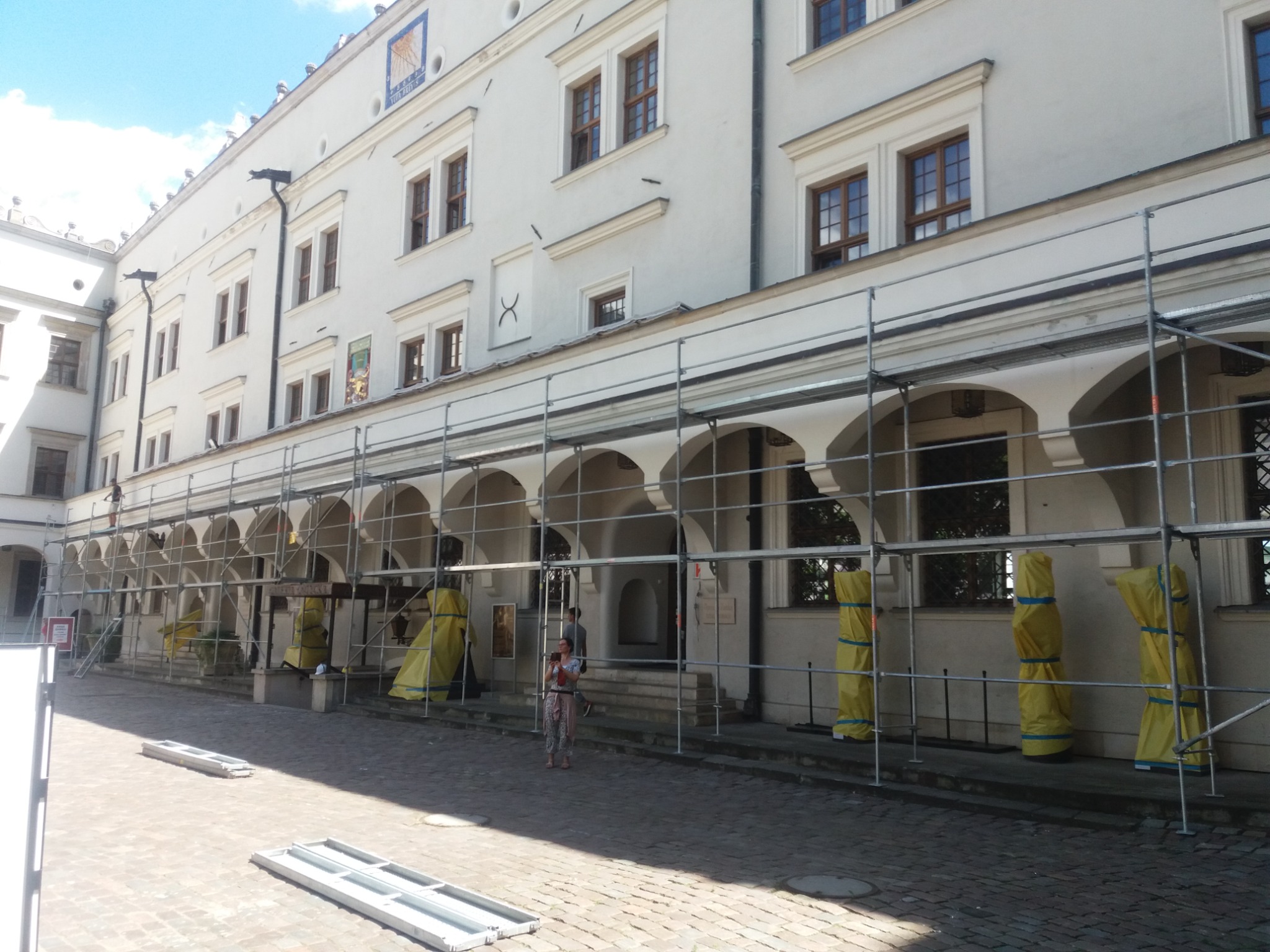 Biały budynek z arkadami w trakcie prac renowacyjnych, osłonięte filary, rusztowania, kobieta z dzieckiem w tle