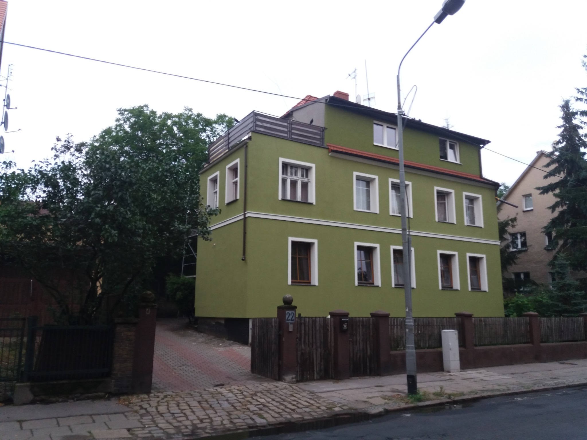 Odnowiony, dwupiętrowy dom z elewacją w odcieniach zieleni, z białymi ramami okiennymi i ciemnobrązową podmurówką, widoczny z poziomu ulicy w pochmurny dzień.