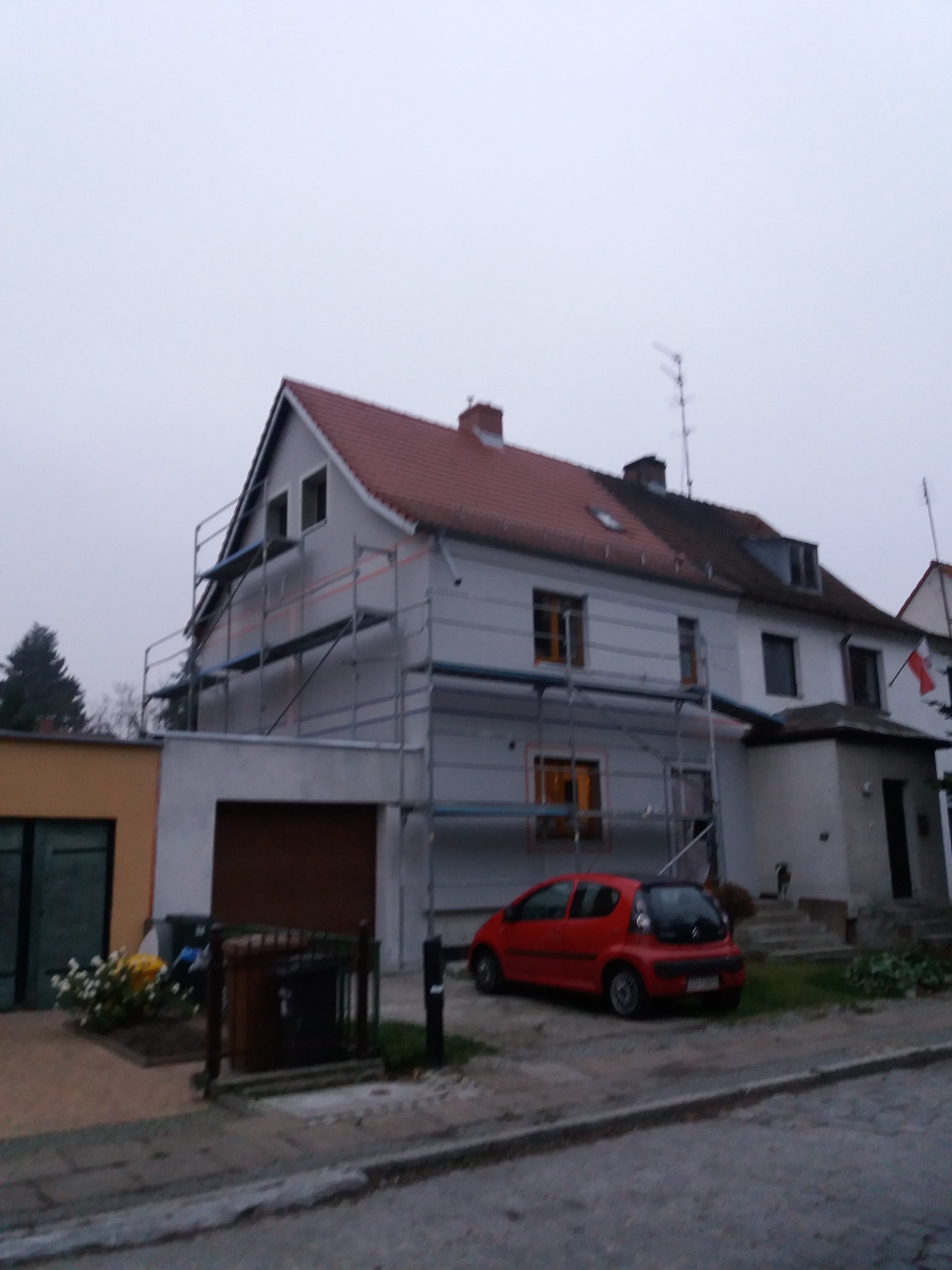 Dwupiętrowy dom w trakcie remontu elewacji, z ustawionymi rusztowaniami i częściowo pomalowaną fasadą na szaro, czerwony samochód osobowy zaparkowany przed budynkiem.