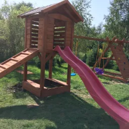 Drewniany plac zabaw z wieżą, zjeżdżalnią i huśtawką na tle zielonego trawnika i drzew, widoczne narzędzia ogrodnicze obok konstrukcji, sugerujące niedawny montaż.