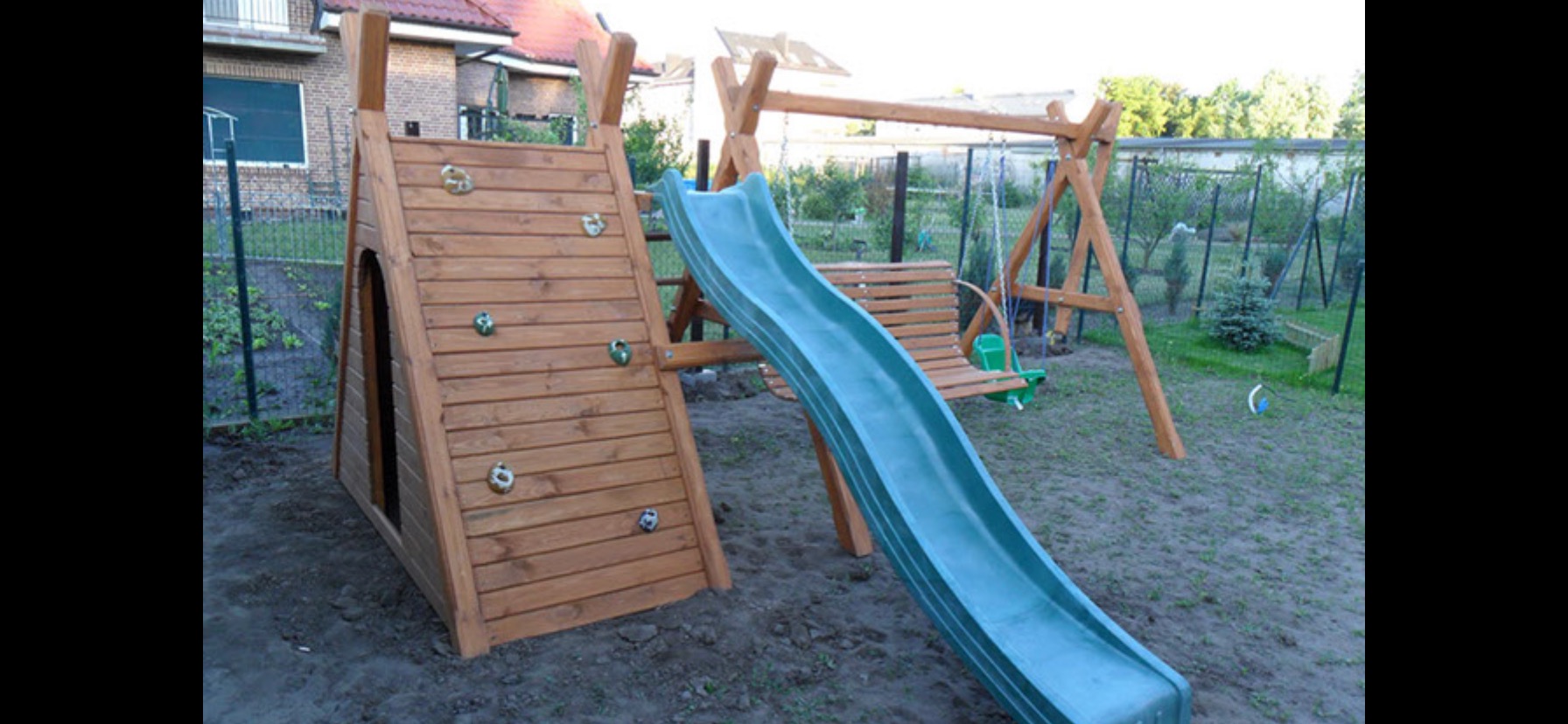 Drewniany plac zabaw z ścianką wspinaczkową, zjeżdżalnią i huśtawką na tle ogrodu z siatką ogrodzeniową.