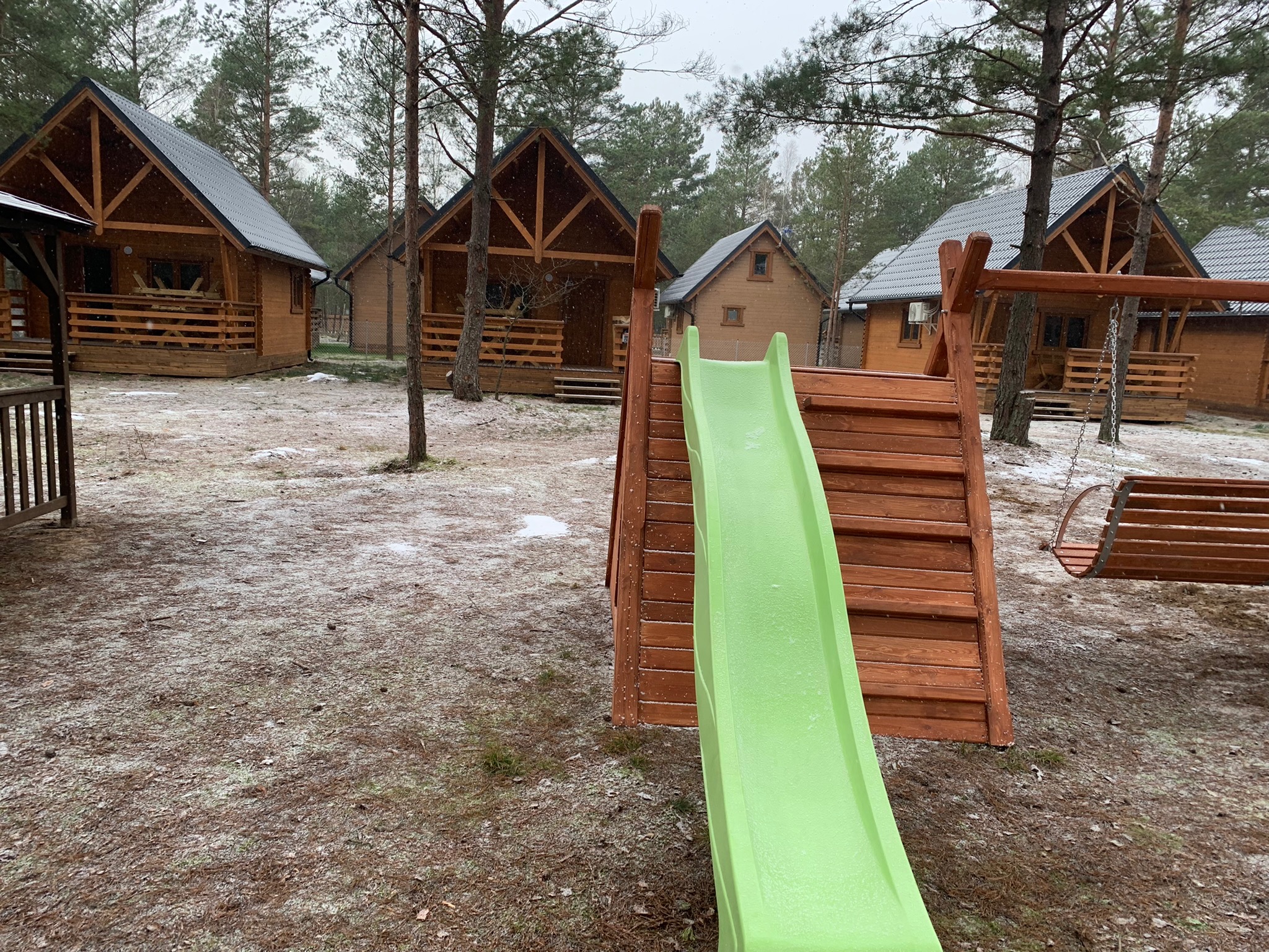 Drewniany plac zabaw z zieloną zjeżdżalnią i huśtawką na tle domków letniskowych w zimowej scenerii.