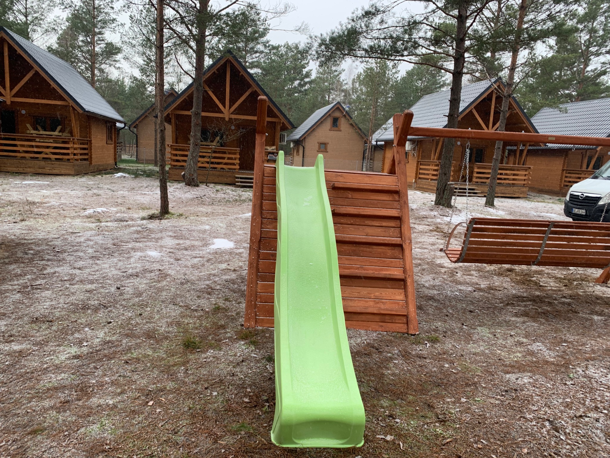 Zielona zjeżdżalnia dla dzieci z drewnianą konstrukcją na placu zabaw, w tle domki letniskowe i lekko ośnieżony teren.