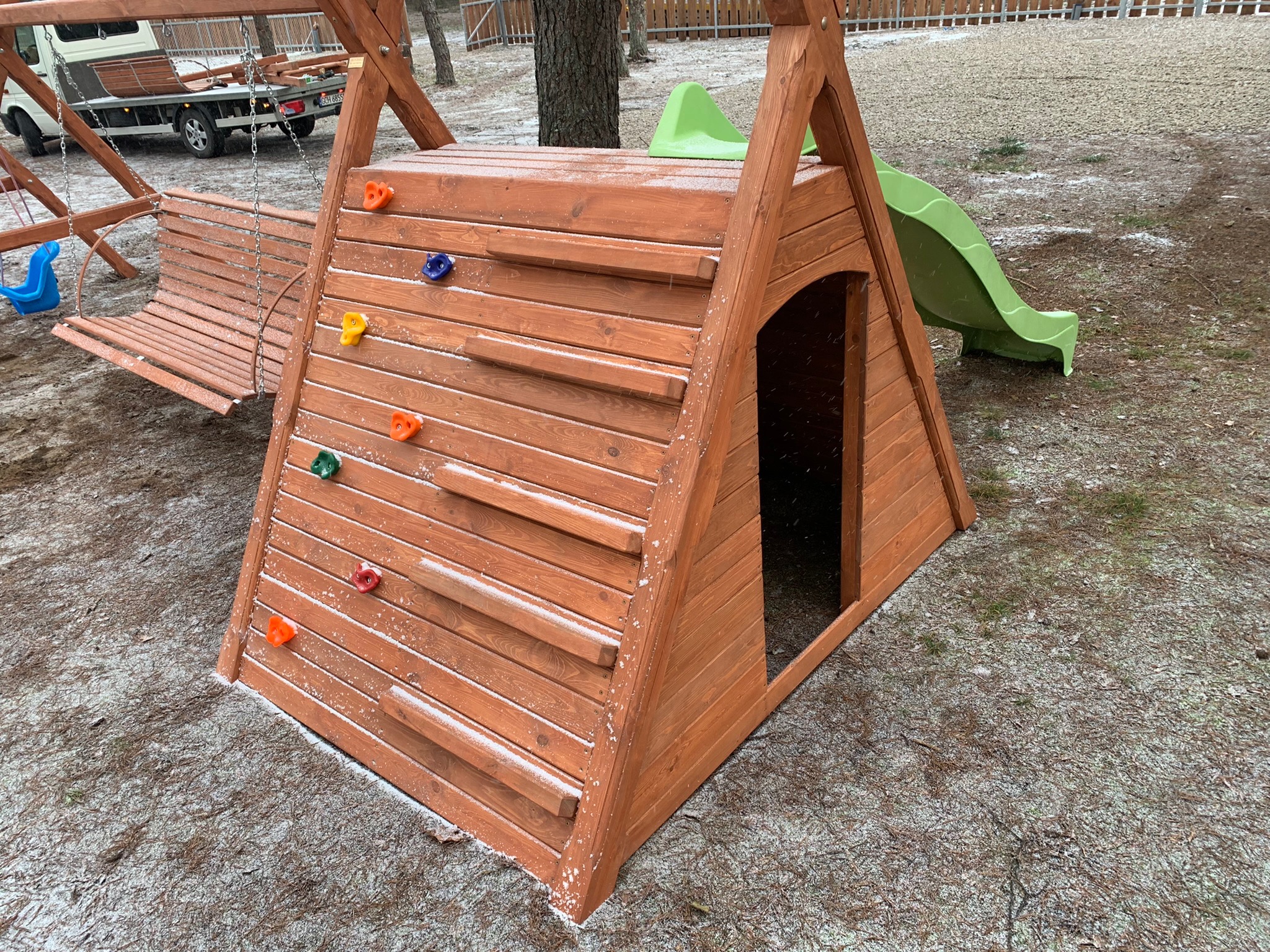 Drewniany plac zabaw z ścianką wspinaczkową z kolorowymi uchwytami i zieloną zjeżdżalnią, lekko przyprószony śniegiem, w tle huśtawka i samochód dostawczy.