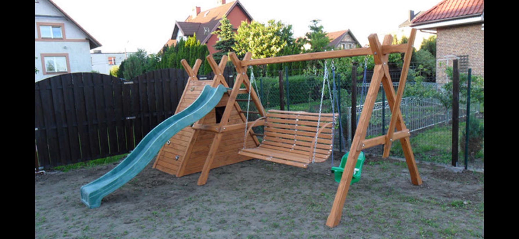 Drewniany plac zabaw z zieloną zjeżdżalnią i huśtawką w ogrodzie, widok na domy w tle.