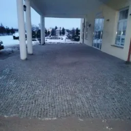 Ułożona kostka brukowa przed budynkiem z kolumnami i oknami, widoczne ślady śniegu i zaparkowany pojazd z urządzeniem.