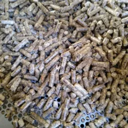 Stos pelletu drzewnego o nieregularnych kształtach i szarej barwie, ułożony na perforowanej metalowej powierzchni.