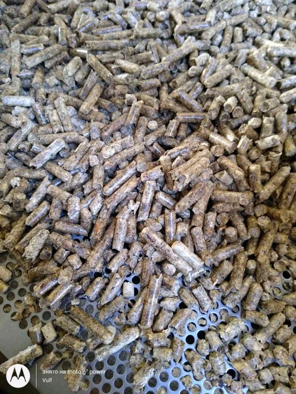 Stos pelletu drzewnego o nieregularnych kształtach i szarej barwie, ułożony na perforowanej metalowej powierzchni.