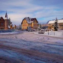 Zimowy krajobraz z żółtym budynkiem hotelu i wieżą z figurką górnika na tle błękitnego nieba.