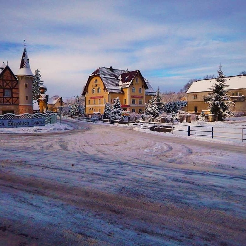 Zimowy krajobraz z żółtym budynkiem hotelu i wieżą z figurką górnika na tle błękitnego nieba.