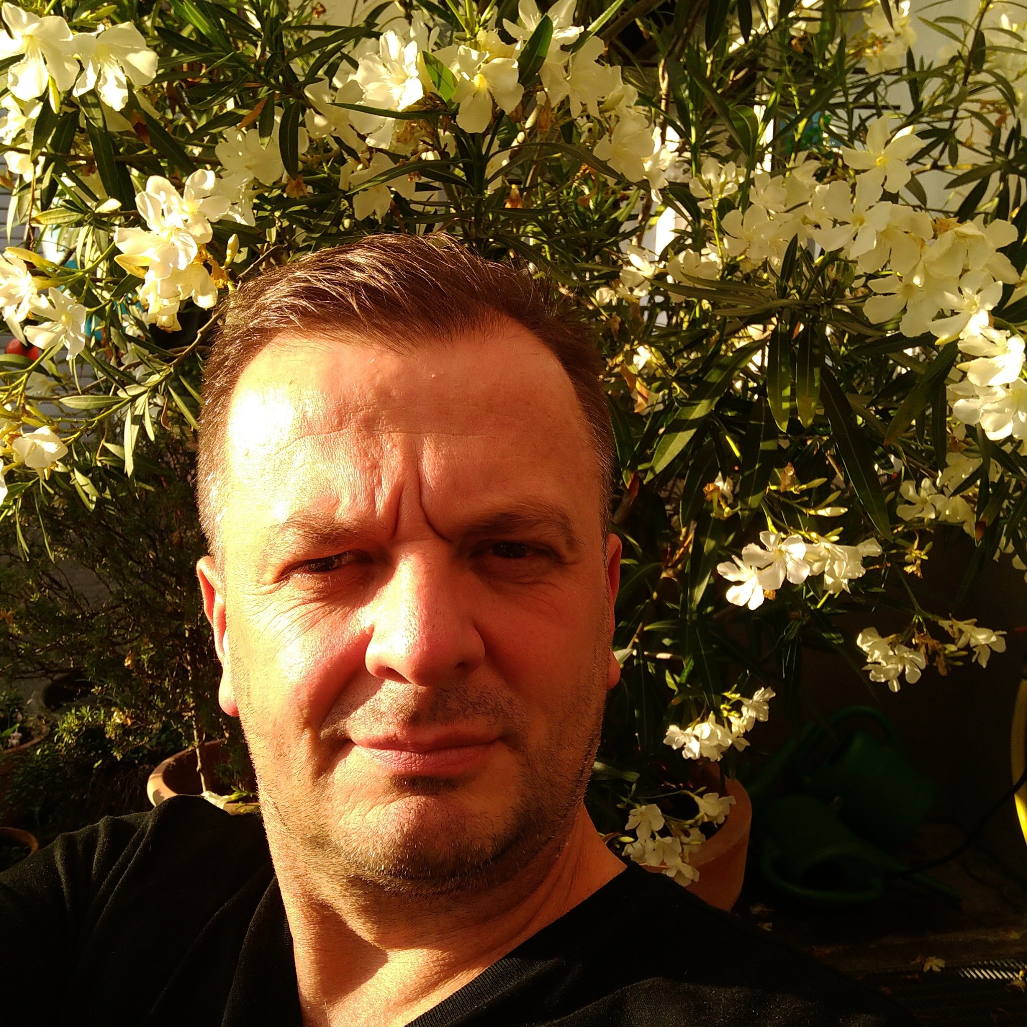 Portret mężczyzny na tle kwitnącego oleandra w doniczce, oświetlonego popołudniowym słońcem.