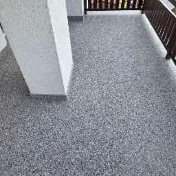 Szara, drobnoziarnista posadzka na balkonie, z widoczną kolumną i brązową balustradą. Widok z góry, ukazujący strukturę powierzchni i elementy architektoniczne.