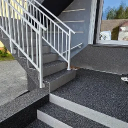 Schody zewnętrzne z szarą, kamyczkową nawierzchnią i białymi barierkami, prowadzące do wejścia z oknem, odbijającym niebo i drzewa.