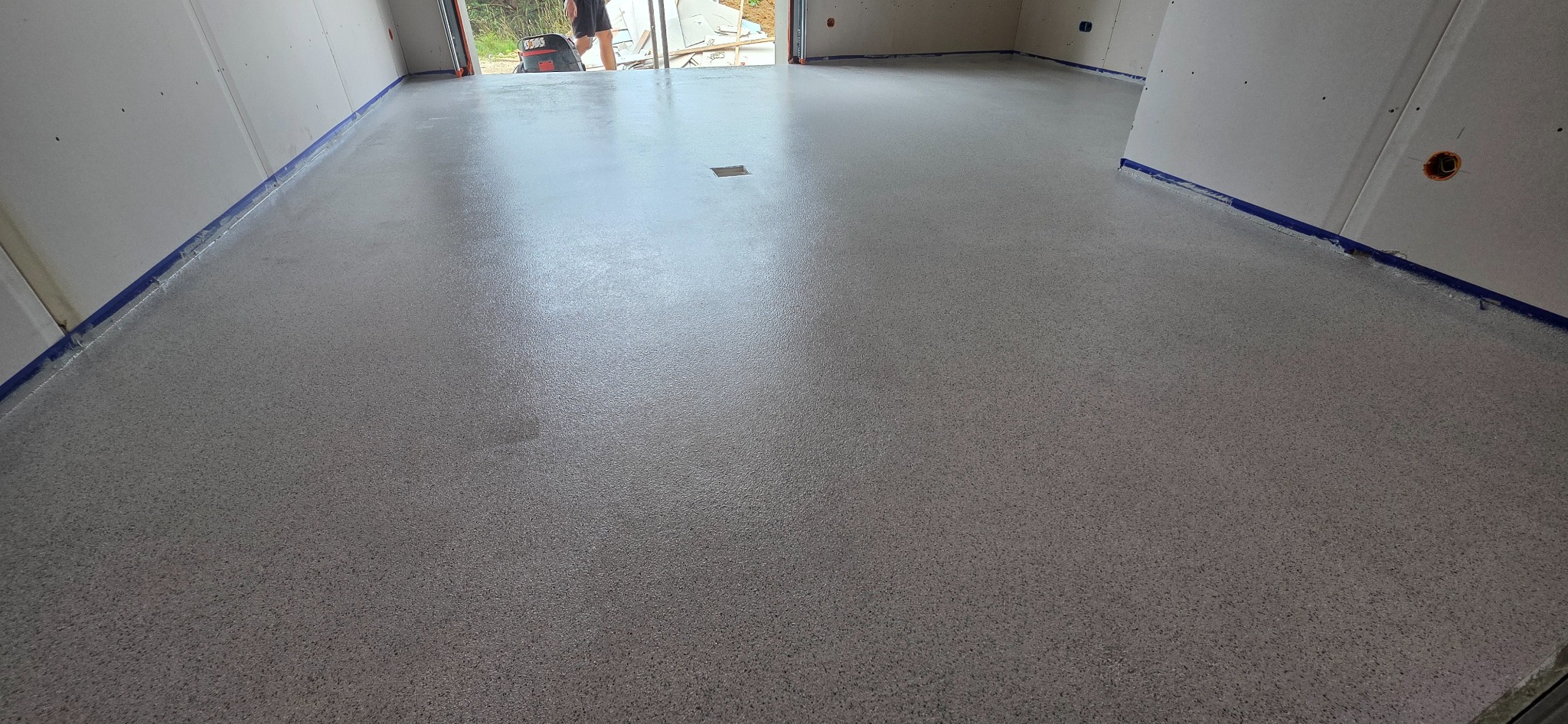 Szara posadzka żywiczna w jasnym pomieszczeniu, zabezpieczona niebieską taśmą malarską przy ścianach. Widoczny otwór w podłodze i osoba w tle.