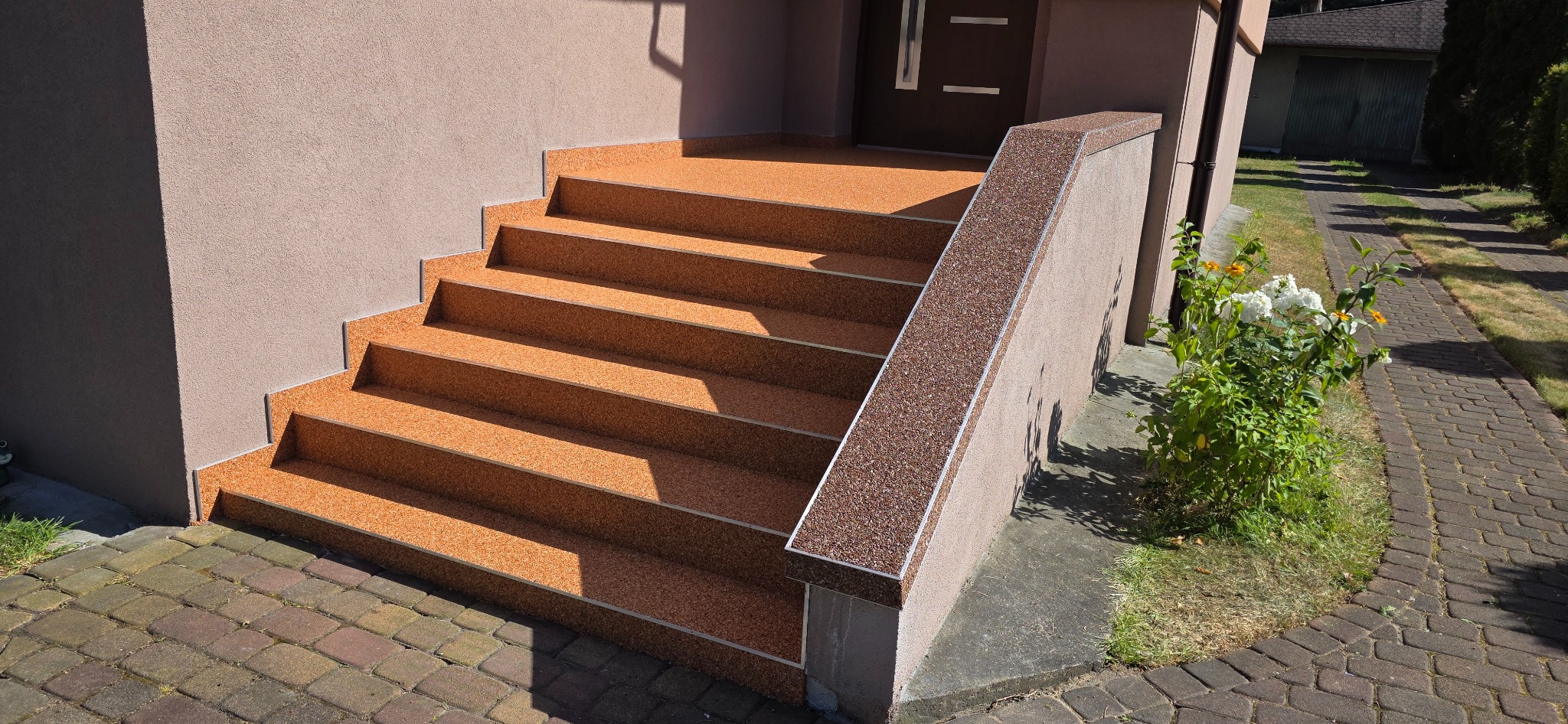 Schody wejściowe do domu wykończone beżową, drobną posadzką żywiczną z metalowymi listwami krawędziowymi. Widok na drzwi wejściowe i fragment chodnika.