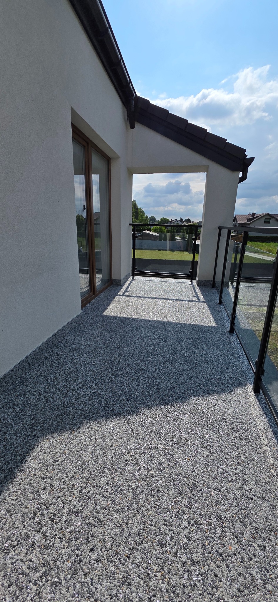Szara posadzka żywiczna na balkonie z przeszkloną balustradą, w tle widok na zieleń i błękitne niebo. Jasne, naturalne światło podkreśla fakturę podłoża.