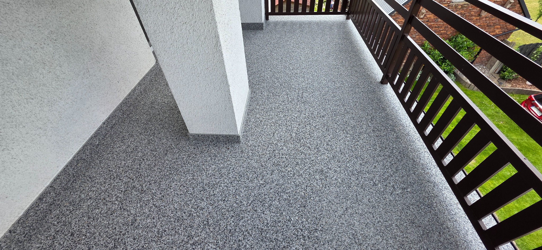 Szara, drobnoziarnista posadzka na balkonie, z widoczną kolumną i brązową balustradą. Widok z góry, ukazujący strukturę powierzchni i elementy architektoniczne.