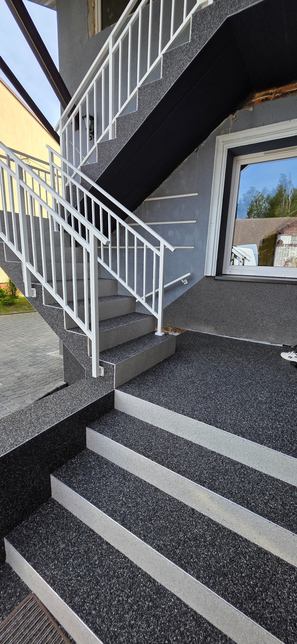Schody zewnętrzne z szarą, kamyczkową nawierzchnią i białymi barierkami, prowadzące do wejścia z oknem, odbijającym niebo i drzewa.
