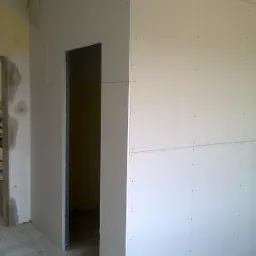 Konstrukcja z płyt kartonowo-gipsowych tworząca ścianę z otworem na drzwi, widoczne ślady po wkrętach, surowy beton na podłodze, fragment instalacji elektrycznej na ścianie.