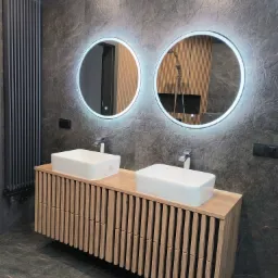 Nowoczesna łazienka z dwoma umywalkami nablatowymi, podświetlanymi okrągłymi lustrami i grzejnikiem żeberkowym na tle ściany z płytek imitujących kamień.