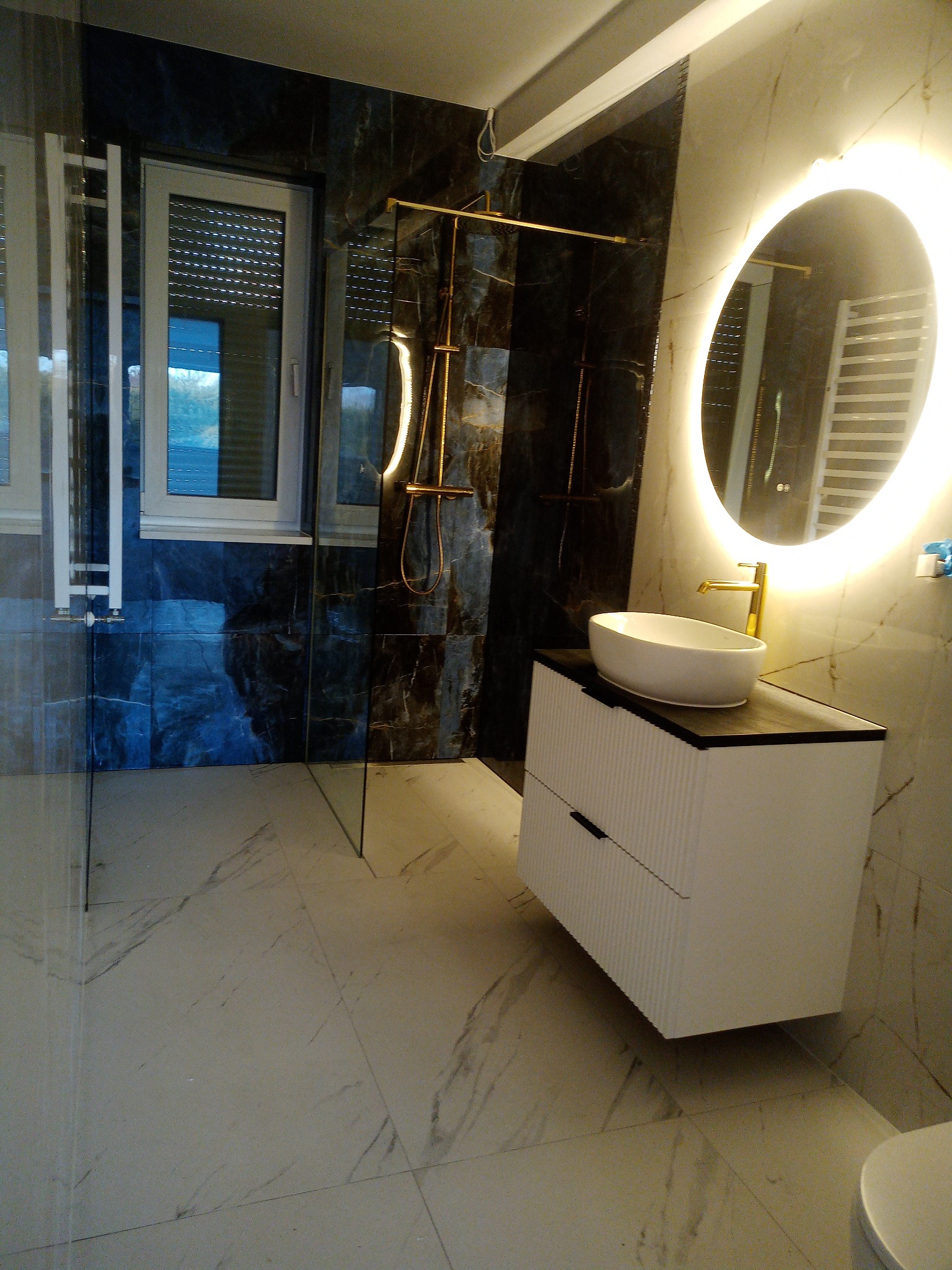 Nowoczesna łazienka z marmurowymi płytkami, owalnym lustrem z podświetleniem LED i białą szafką pod umywalkę z złotą armaturą. Kabina prysznicowa ze złotymi elementami.