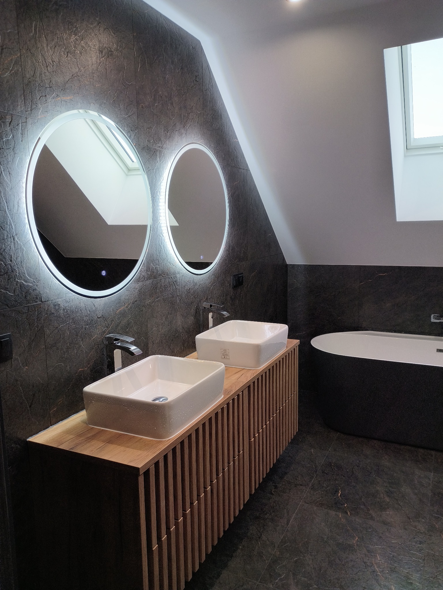 Nowoczesna łazienka na poddaszu z dwoma umywalkami nablatowymi, podświetlanymi lustrami i ciemną armaturą. Uwagę zwraca drewniana szafka pod umywalki i grafitowa glazura.