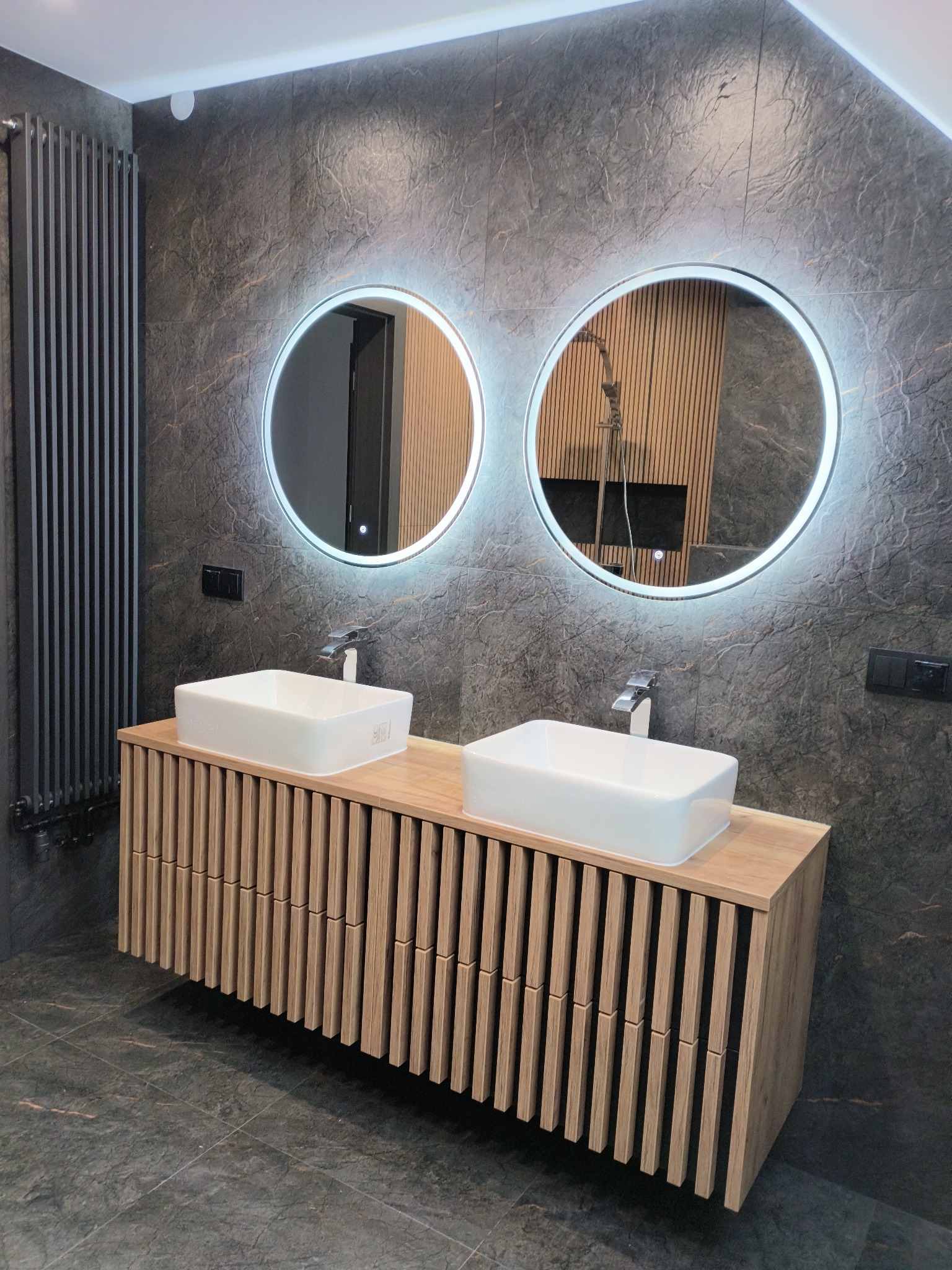 Nowoczesna łazienka z dwoma umywalkami nablatowymi, podświetlanymi okrągłymi lustrami i grzejnikiem żeberkowym na tle ściany z płytek imitujących kamień.