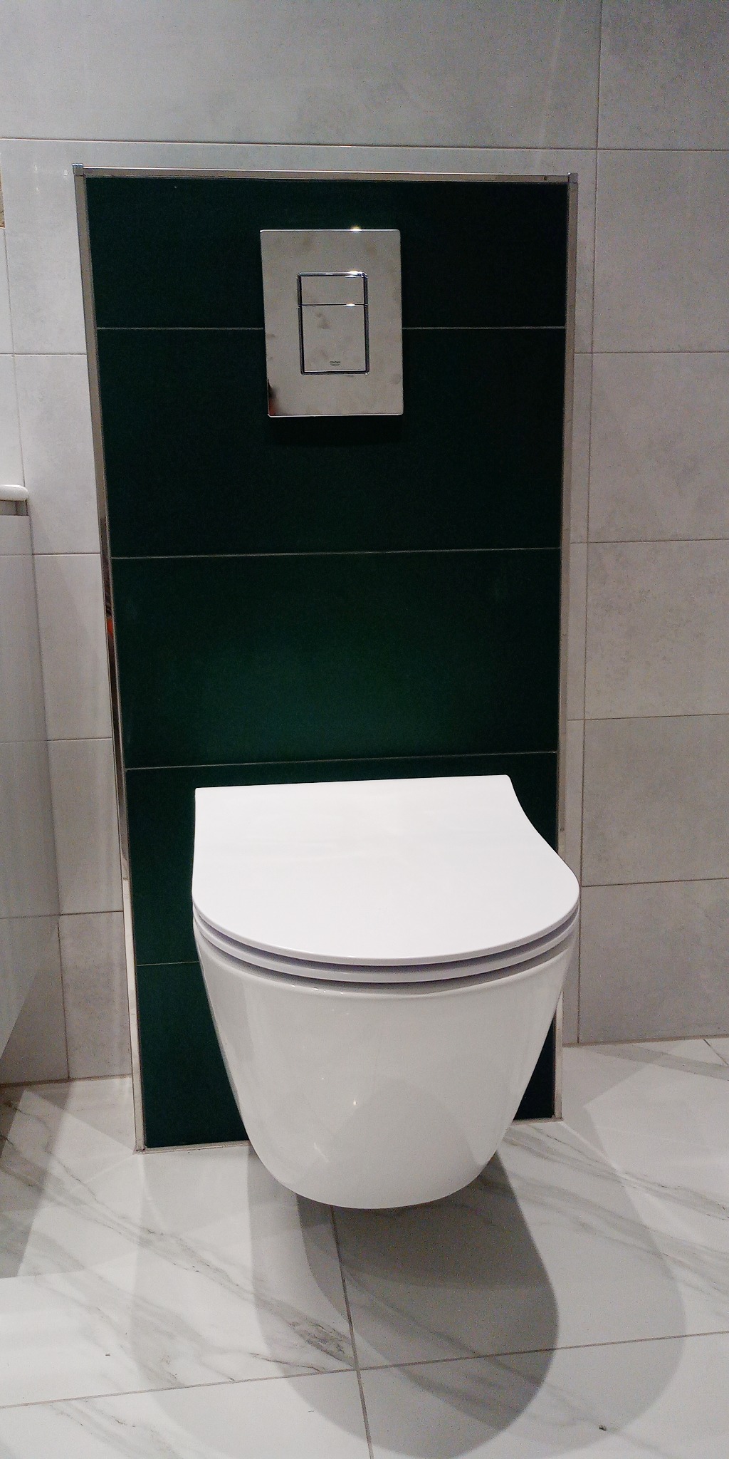 Wisząca toaleta z chromowanym przyciskiem spłukującym na tle ciemnozielonych płytek, otoczona jasnymi płytkami imitującymi marmur.